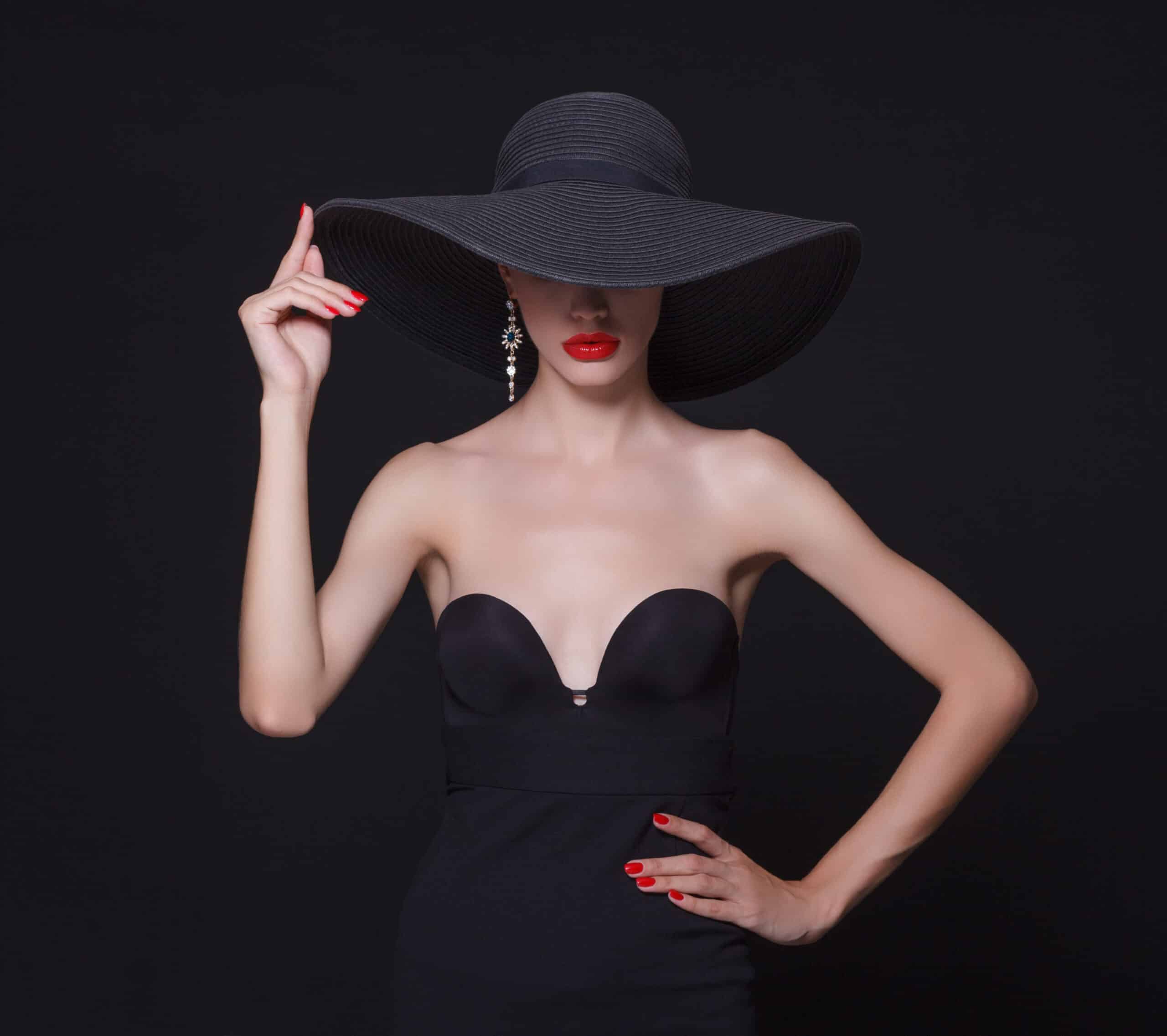 Femme élégante en robe bustier noire, grand chapeau et rouge à lèvres éclatant.