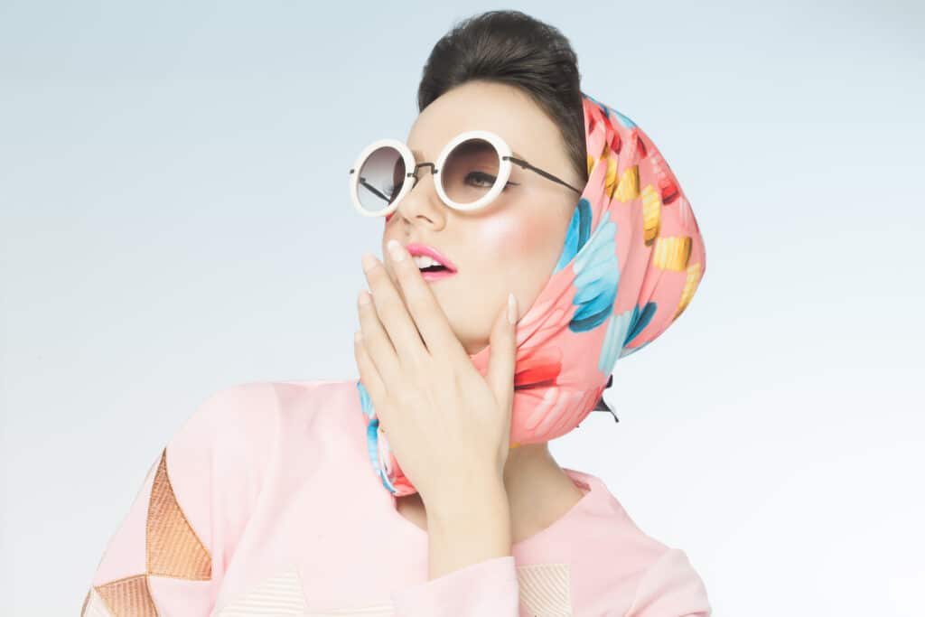 Look Rétro Vintage : Femme avec Lunettes et Foulard Rose Femme élégante style vintage, lunettes rondes blanches, foulard floral rose, main sur la bouche, look rétro chic.