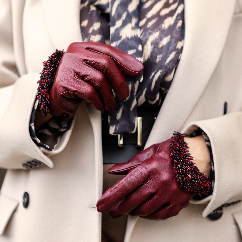 Gants en Cuir Bordeaux avec Poignets Perlés Élégants Gants en cuir bordeaux avec poignets perlés rouges et noirs, portés avec un manteau beige et une écharpe à motifs.
