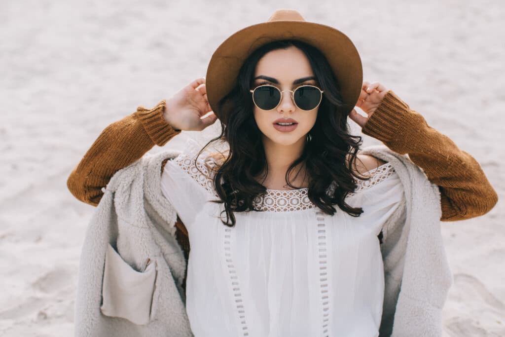 Mode Bohème Chic : Femme avec Chapeau et Lunettes de Soleil Femme brune avec chapeau marron et lunettes rondes, style bohème chic sur une plage de sable blanc.