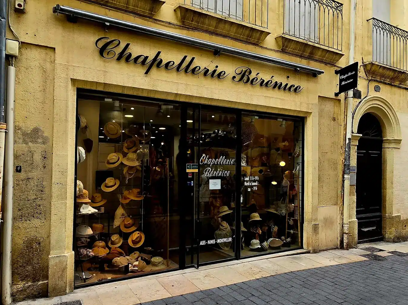 Boutique Chapellerie Bérénice avec vitrine de chapeaux variés et façade traditionnelle en pierre jaune.