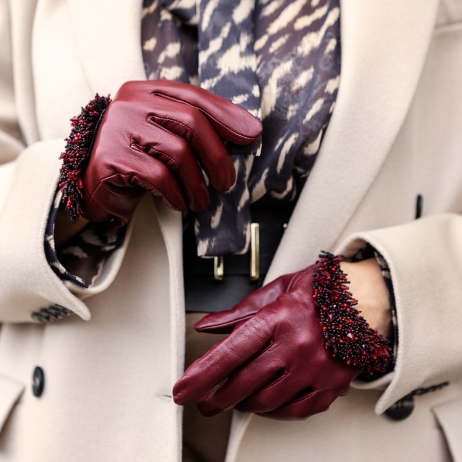 Gants en Cuir Bordeaux et Perles : Élégance Hivernale Gants en cuir bordeaux avec poignets perlés, portés avec un manteau beige élégant et une écharpe imprimée.