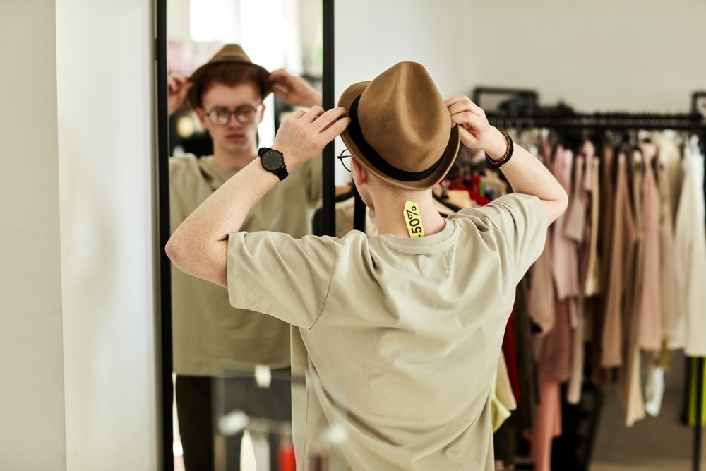 Homme essayant un chapeau fedora marron en soldes -50% devant un miroir dans une boutique de vêtements.