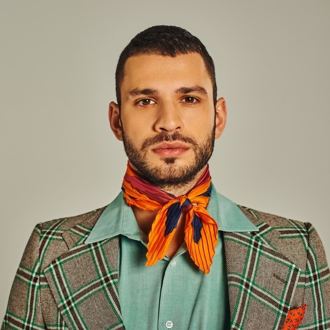 Look Homme Élégant : Blazer Carreaux et Foulard Orange Homme barbu en blazer à carreaux, chemise turquoise et foulard orange noué, regardant fixement l'objectif.